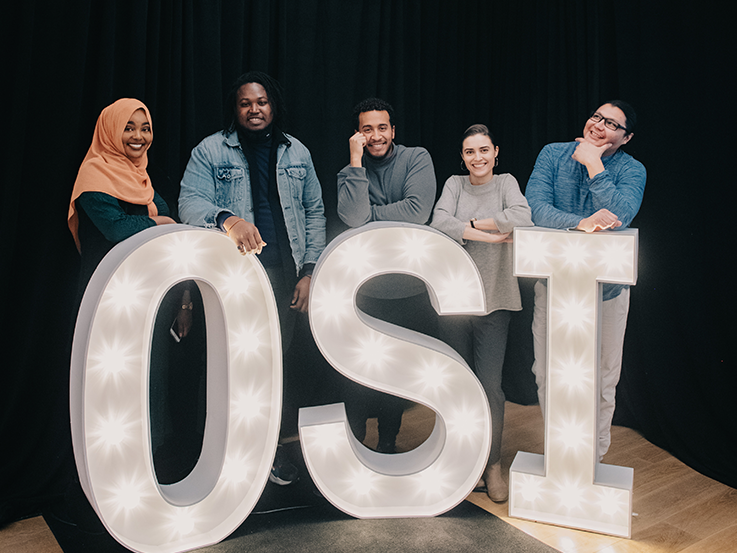 a group of 4 students stand leaning behind 3 large letter lights spelling OSI